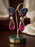 A pair of dramatic teardrop dangle earrings displayed on a gold jewelry stand with dark velvet cushions. The earrings feature large faceted magenta-pink teardrop crystals, a small round clear stone, and a silver-toned setting.
