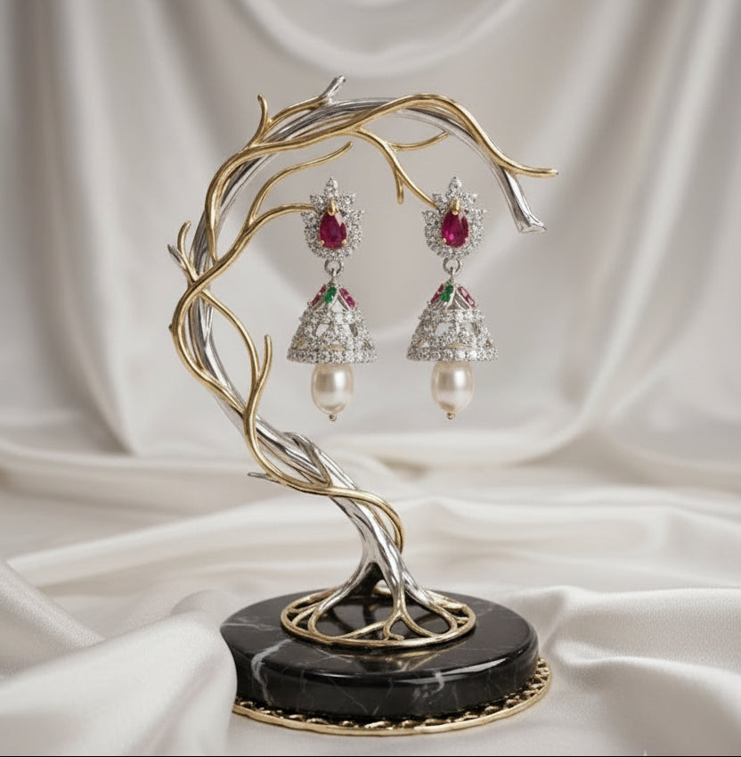 A pair of ornate dangling earrings displayed on a sculptural branch-shaped stand. The earrings feature silver-toned bell-shaped drops set with clear stones, a central red gemstone, and a white pearl at the bottom, mounted on a black marble base in an elegant indoor setting.