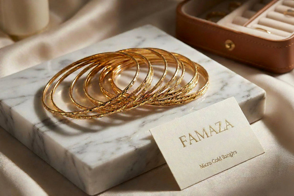 A high-end lifestyle photograph of 12 sleek, micro gold-plated copper bangles with a diamond-cut texture, displayed on a white marble block next to an Aurum Collective Micro Gold Bangles card.