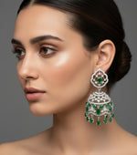 A pair of silver finish Jhumka earrings with emerald green stones and American diamond accents displayed on an earrings stand.