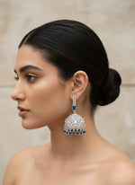 A pair of silver finish Jhumka earrings with American diamonds and blue sapphire stone details, displayed against a plant backdrop.