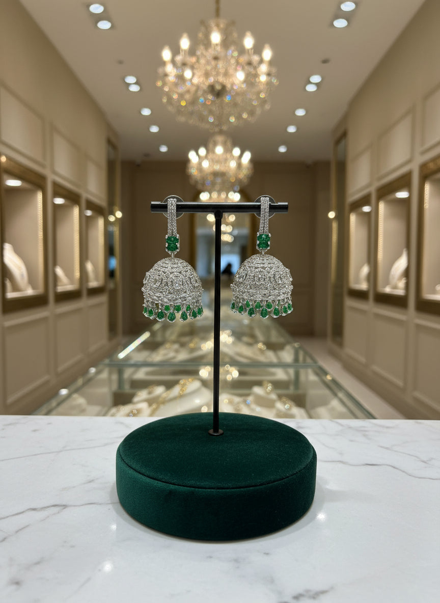 A pair of silver finish Jhumka earrings with American diamonds and green stone detailing, displayed against a neutral background.