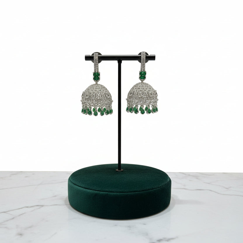 A pair of silver finish Jhumka earrings with American diamonds and green stone detailing, displayed against a neutral background.