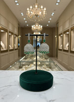 A pair of silver finish Jhumka earrings with American diamonds and green stone detailing, displayed against a neutral background.