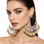 A close-up of a woman wearing large ornate silver hoop earrings with dangling gold coin charms and white bead accents against a clean white background.