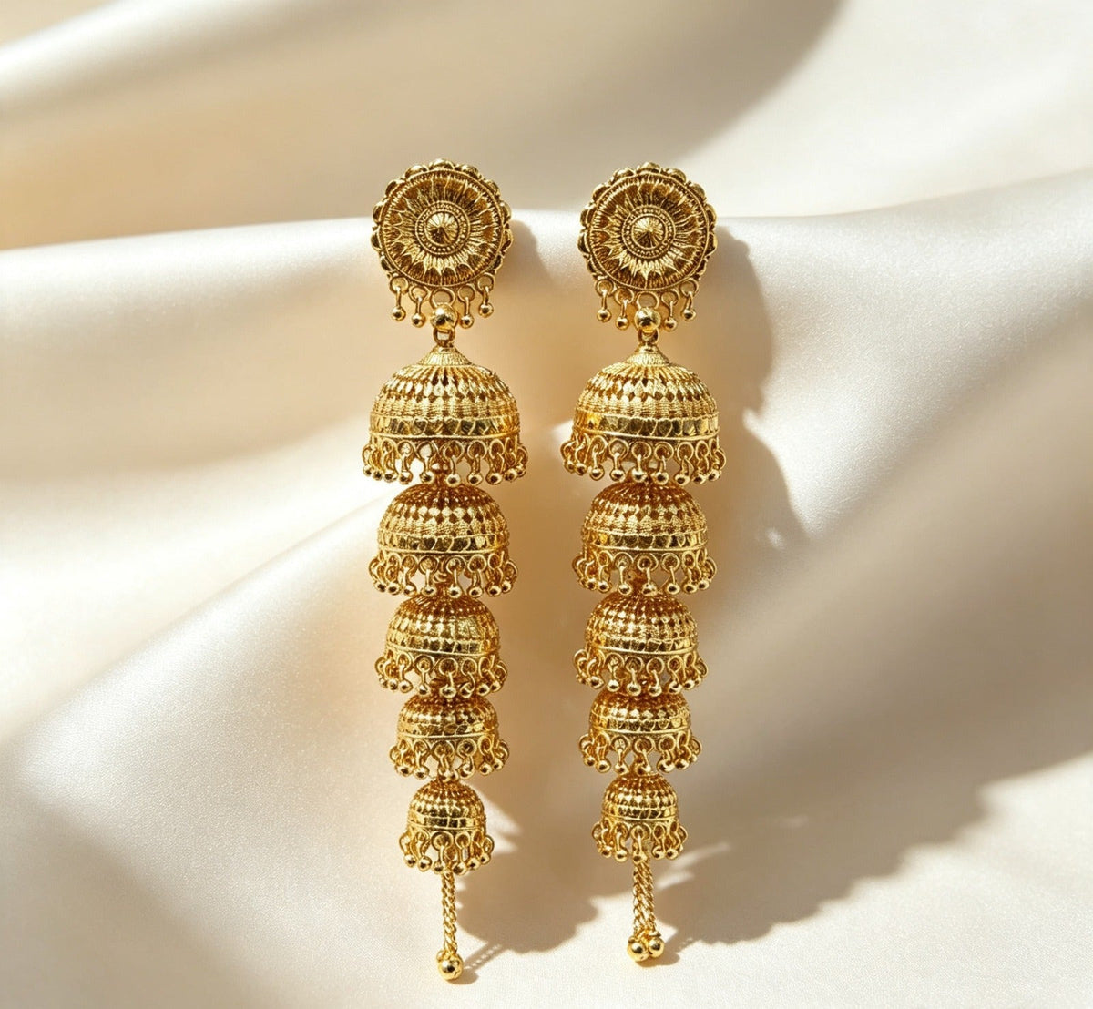 Pair of 5-tier cascading gold jhumka earrings with filigree rosette tops and five descending domed bells, handcrafted gold-plated traditional South Asian bridal statement jewelry on cream satin background.