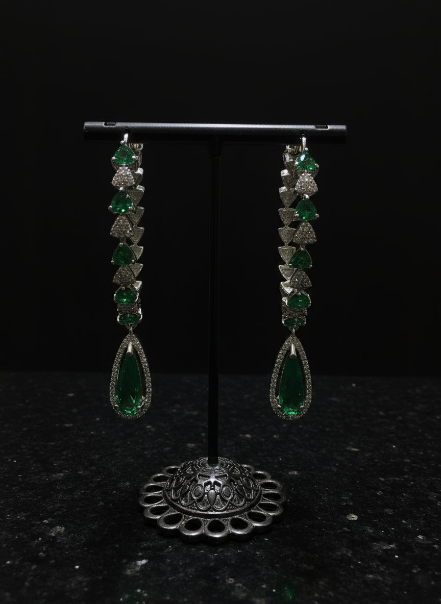 A pair of silver finish American diamond Jhumka bali earrings with green and clear stones on a stand.
