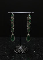 A pair of silver finish American diamond Jhumka bali earrings with green and clear stones on a stand.