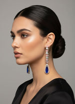 A pair of silver finish American diamond Jhumka bali earrings with green and clear stones on a stand.