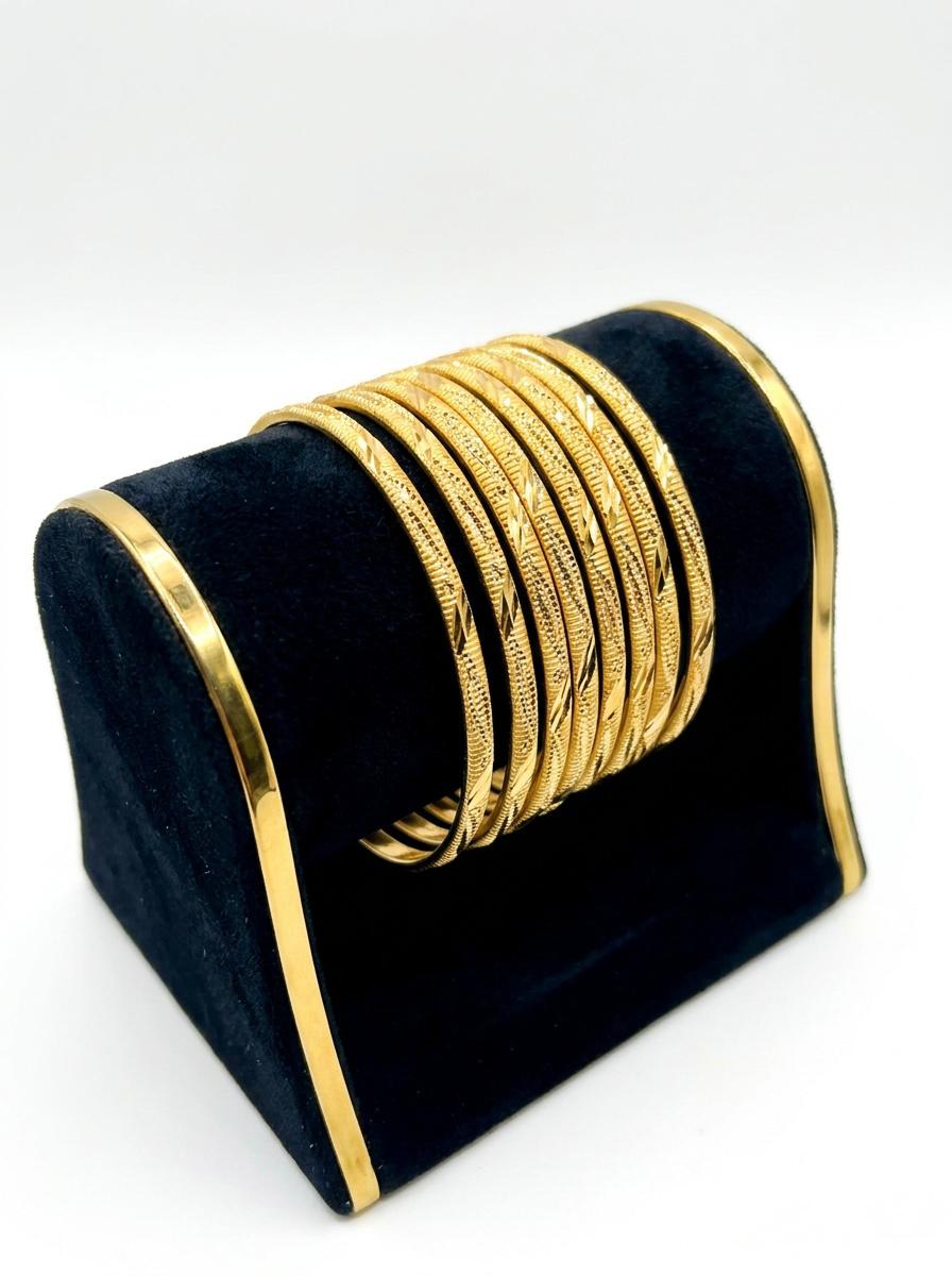 A set of eight micro gold-plated copper bangles with an intricate twisted rope and wavy vine texture, displayed on a posh black velvet and gold-trimmed jewelry stand against a white background.