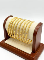 A set of eight micro gold-plated copper bangles featuring an intricate twisted rope and wavy vine texture, displayed on a posh wooden and cream suede jewelry stand against a white background.