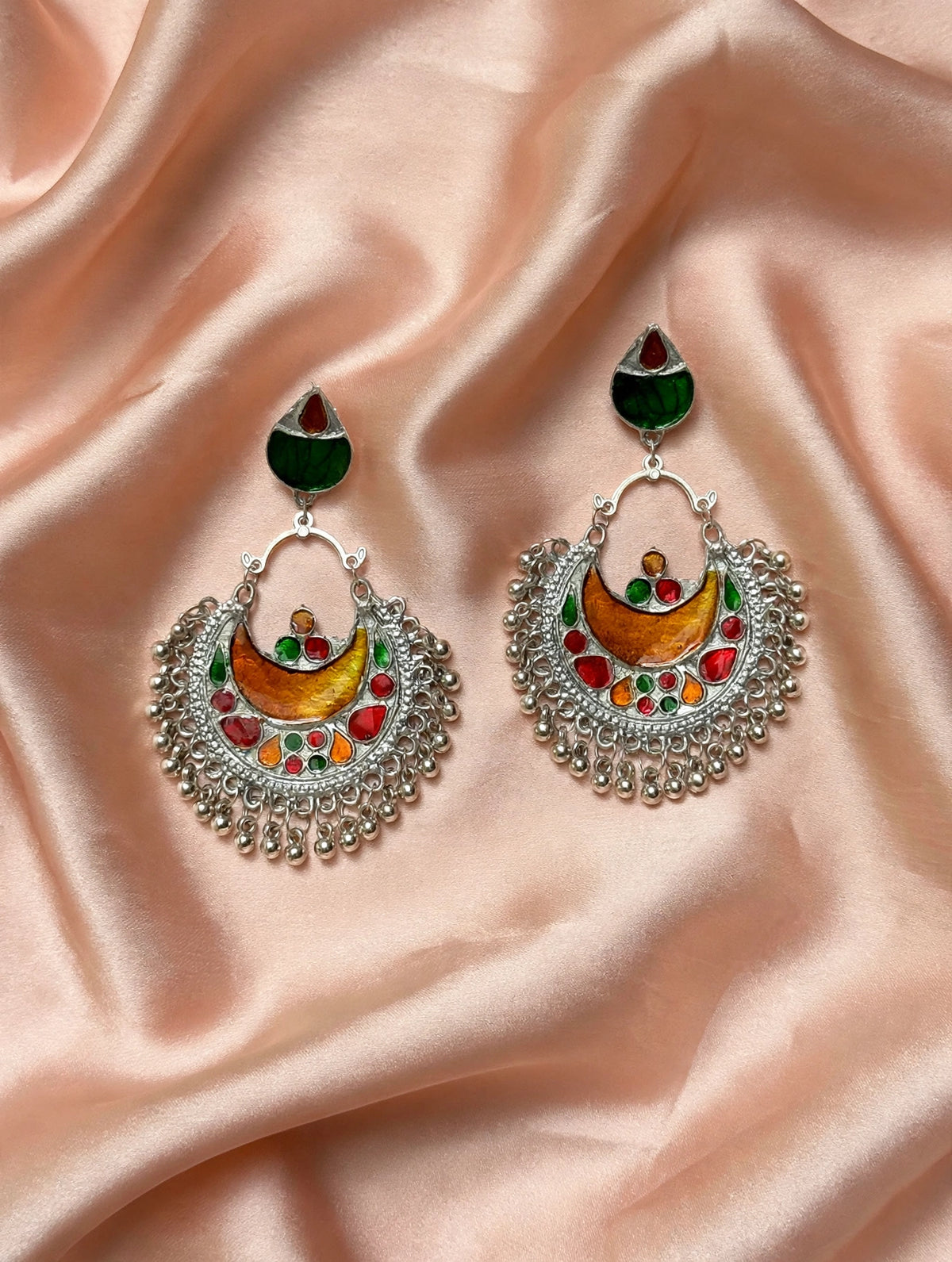 A pair of ornate silver-toned Indian-style dangle earrings with colorful enamel detailing, displayed on a draped peach satin fabric background.