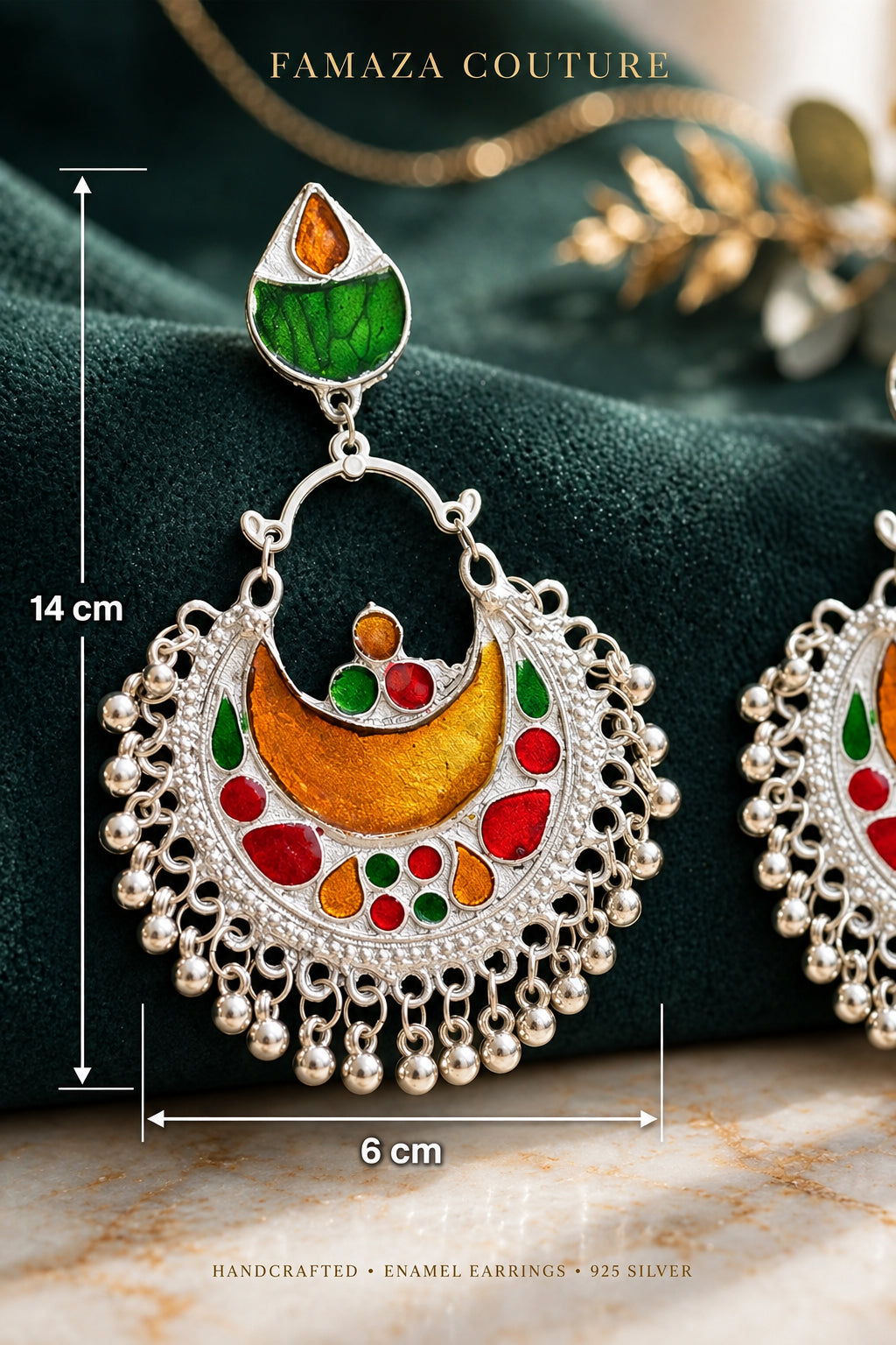 A close-up of a silver crescent-shaped earring with colorful enamel details in green, orange, red, and yellow, displayed on a dark green velvet surface with other matching earrings partially visible.