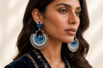A close-up lifestyle shot of a woman wearing ornate silver-toned crescent-shaped dangle earrings with vibrant blue enamel and beadwork. The brand name 'TAMAZA COUTURE' is visible at the bottom.