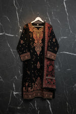 A black embroidered tunic top with red and gold patterns, hanging on a rack between blue and green patterned dresses.