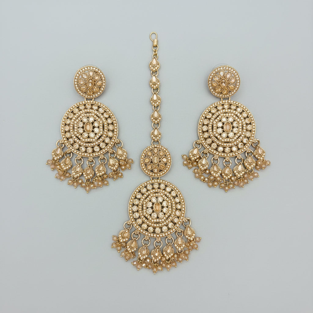 A Rose Gold-toned Indian bridal jewellery set featuring a maang tikka and twodangle earrings all embellished with intricate crystal work and hanging tassels, displayed on a black posh velvet surface.