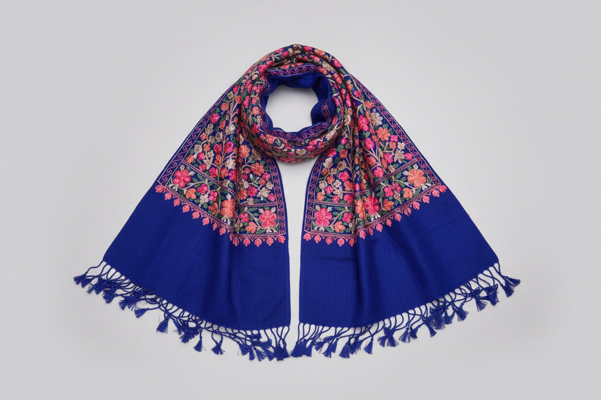 Ablue scarf with elaborate pink and green floral aari embroidery and tassels on the ends, displayed on a dark background.