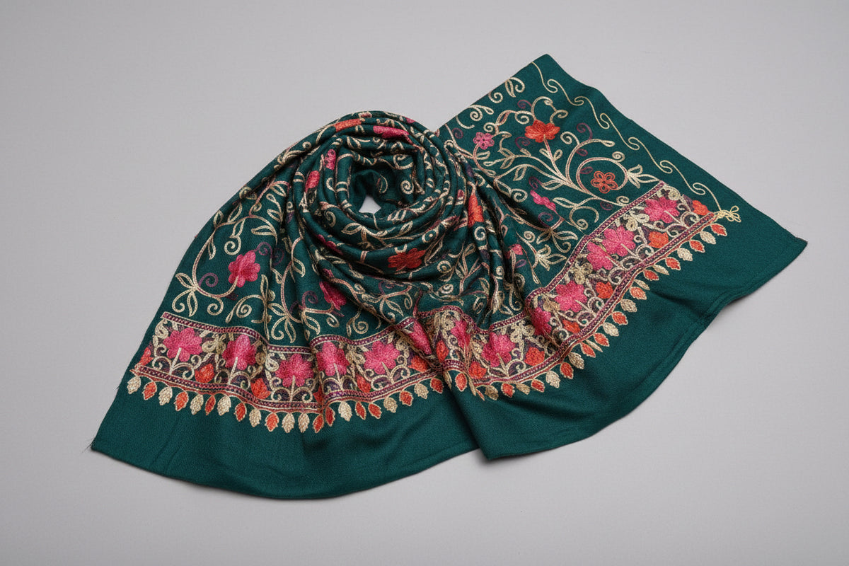 A bottle green Kashmiri embroidered pashmina scarf with tassels hanging at the ends, displayed on a wooden surface.