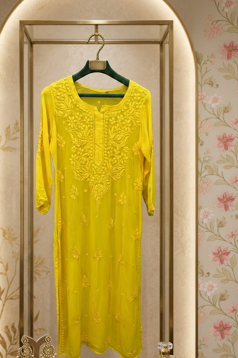 A bright yellow embroidered kurti with three-quarter sleeves, hanging on a green hanger inside a glass display rack. A folded matching yellow fabric piece and a small perfume bottle sit on the wooden shelf below.