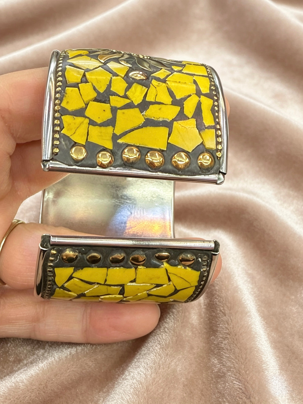 A close-up of a hand holding a wide cuff bracelet with a yellow mosaic tile pattern and gold-toned metal details, set against a draped champagne satin fabric background.
