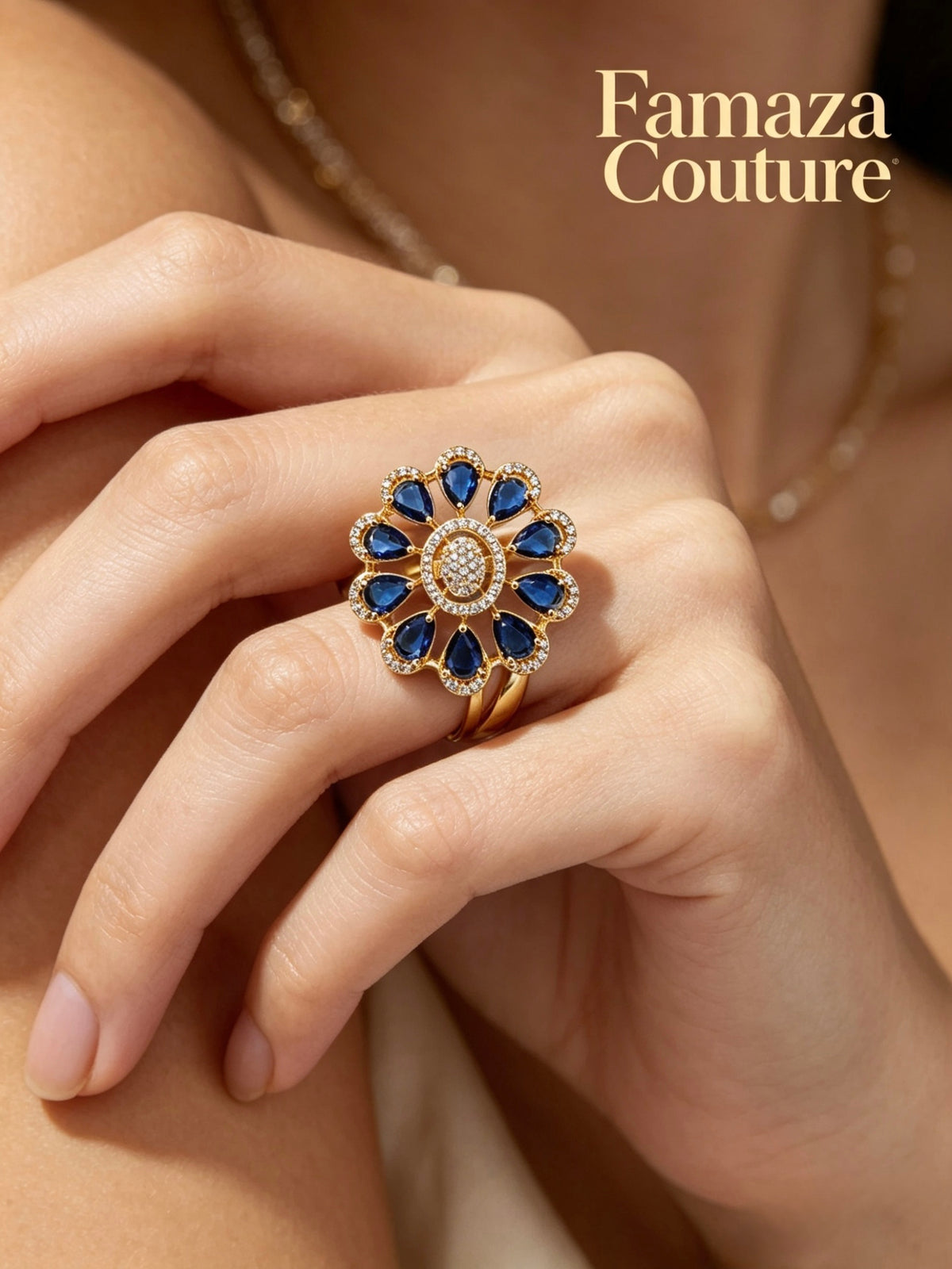 A close-up of a hand wearing a gold flower-shaped ring with deep blue gemstones and a central cluster of clear stones. The image has the brand name 'Famaza Couture' printed in gold text in the upper right corner.