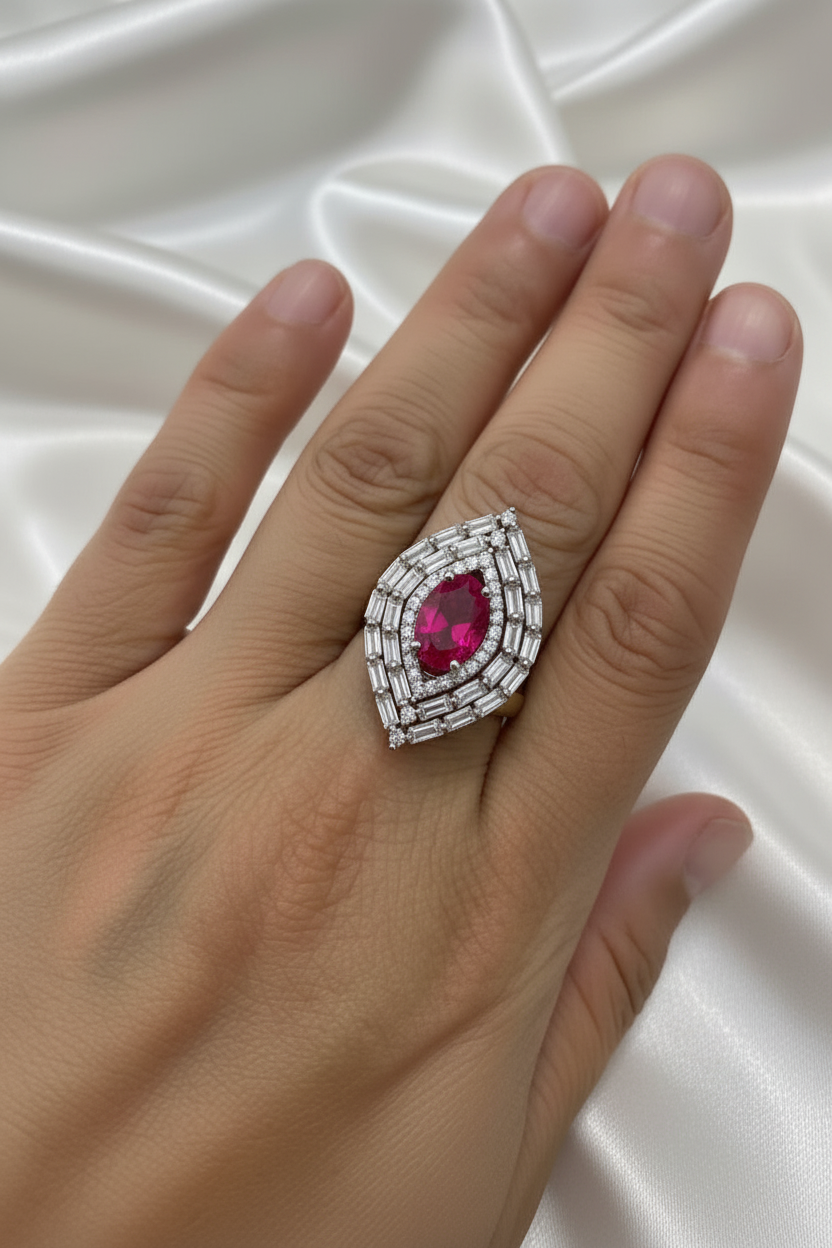 A close-up of a hand wearing a large statement ring with a central marquise-cut pink gemstone surrounded by silver-toned layered bands set with small clear stones, photographed against draped white satin fabric.