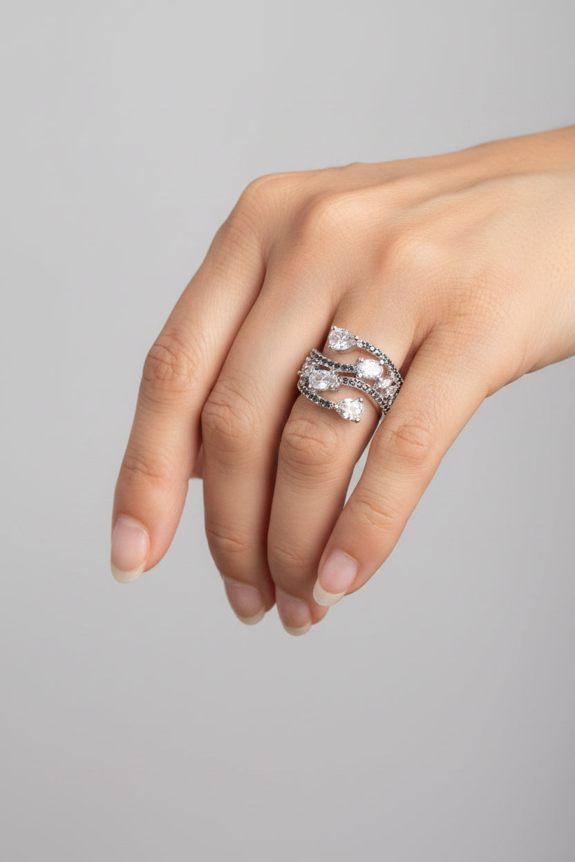 A close-up of a hand wearing a silver-toned multi-band ring set with clear heart-shaped and round gemstones, resting on a black surface.