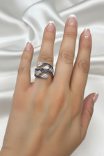 A close-up of a hand with a French manicure wearing a multi-band ring. The ring features silver-toned bands set with round clear stones and rows of small dark blue stones, photographed against a draped white satin fabric background.