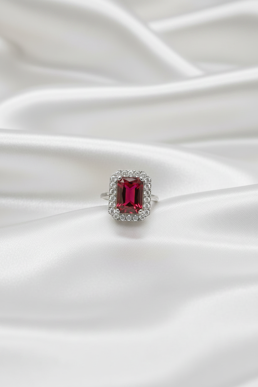 A silver ring featuring a rectangular emerald-cut red gemstone surrounded by a halo of small clear stones, resting on draped white satin fabric.