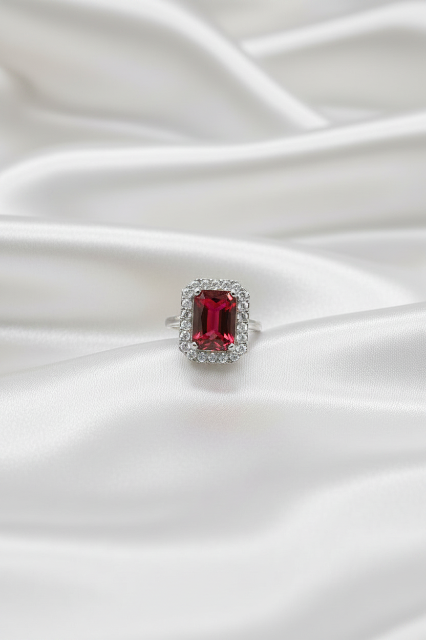 A silver ring featuring a rectangular emerald-cut red gemstone surrounded by a halo of small clear stones, resting on draped white satin fabric.