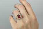 A close-up of a hand wearing a yellow gold ring with a rectangular red gemstone surrounded by a halo of small clear stones, set against a dark blue background.