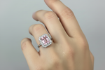 A close-up of a hand wearing a silver ring featuring a large rectangular pink gemstone surrounded by a halo of small clear stones.