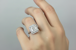 A close-up of a hand wearing a silver-toned ring with a large rectangular baguette-cut center stone surrounded by smaller stones in a halo setting, against a soft light gray background.