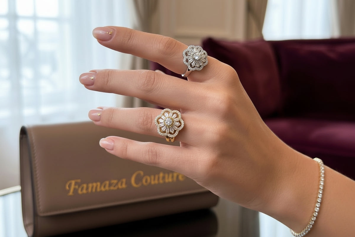 A close-up of a hand wearing two large flower-shaped rings, one silver-toned and one gold-toned, while holding a glass of champagne. A matching pearl bracelet is also visible, with a brown leather clutch and a burgundy sofa in the background.