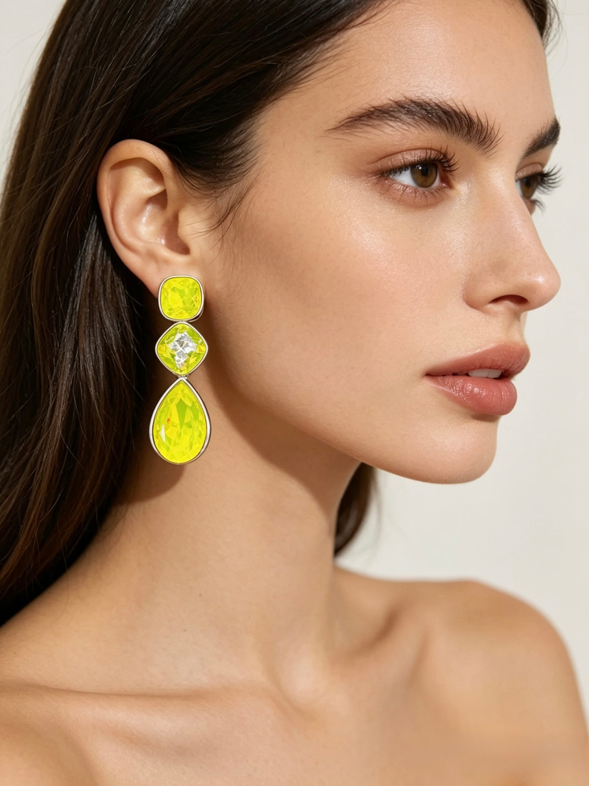 A close-up of a model wearing a vibrant yellow three-stone drop earring with clear gemstones in a gold-tone setting, against a clean, light beige background.