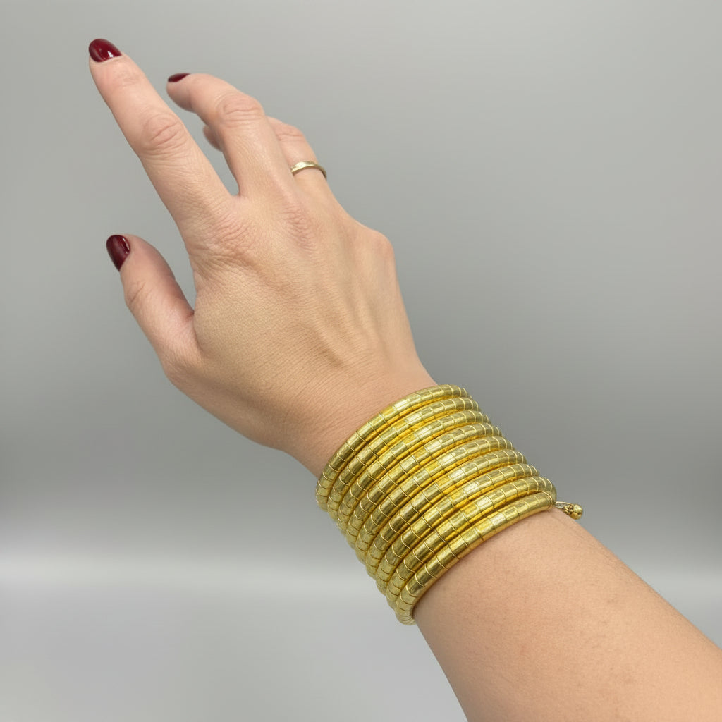 A close-up of a person's hand and wrist wearing a stack of wide, rigid gold bangles against a dark textured background.
