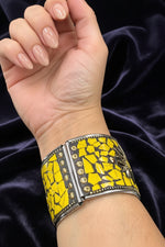A close-up of a person's wrist wearing a wide cuff bracelet with a yellow and black mosaic tile pattern and gold-tone studs, set against a dark purple velvet background.