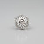 A close-up of a silver-toned flower-shaped ring with a central round diamond and surrounding baguette and round diamonds, held against a dark display cone with fingers visible in the background.