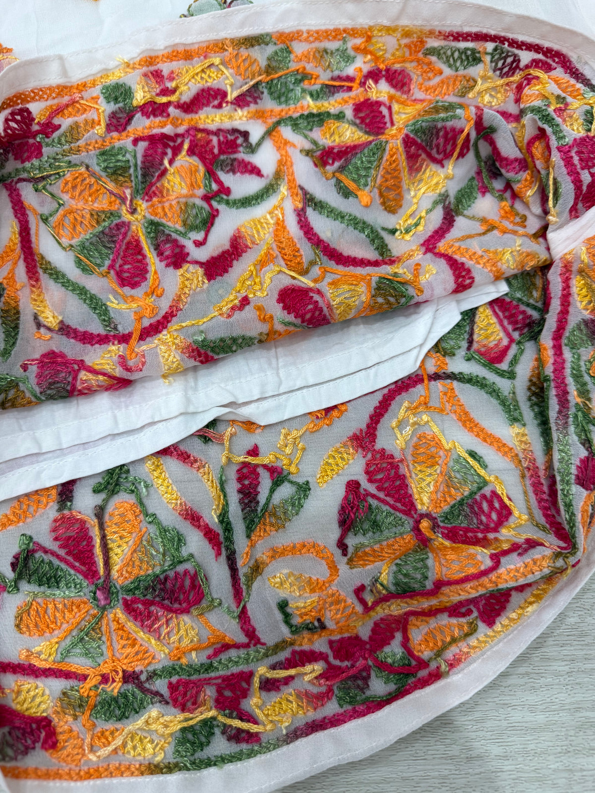 A close-up of a white embroidered fabric, likely a dupatta or scarf, featuring intricate floral and paisley embroidery in vibrant orange, yellow, green, and magenta pink.
