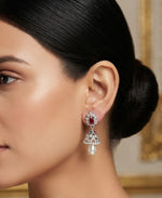 A close-up of a woman wearing a dangling chandelier earring with a red ruby stone, clear pavé setting, and a white pearl drop, shown against a softly blurred interior background.