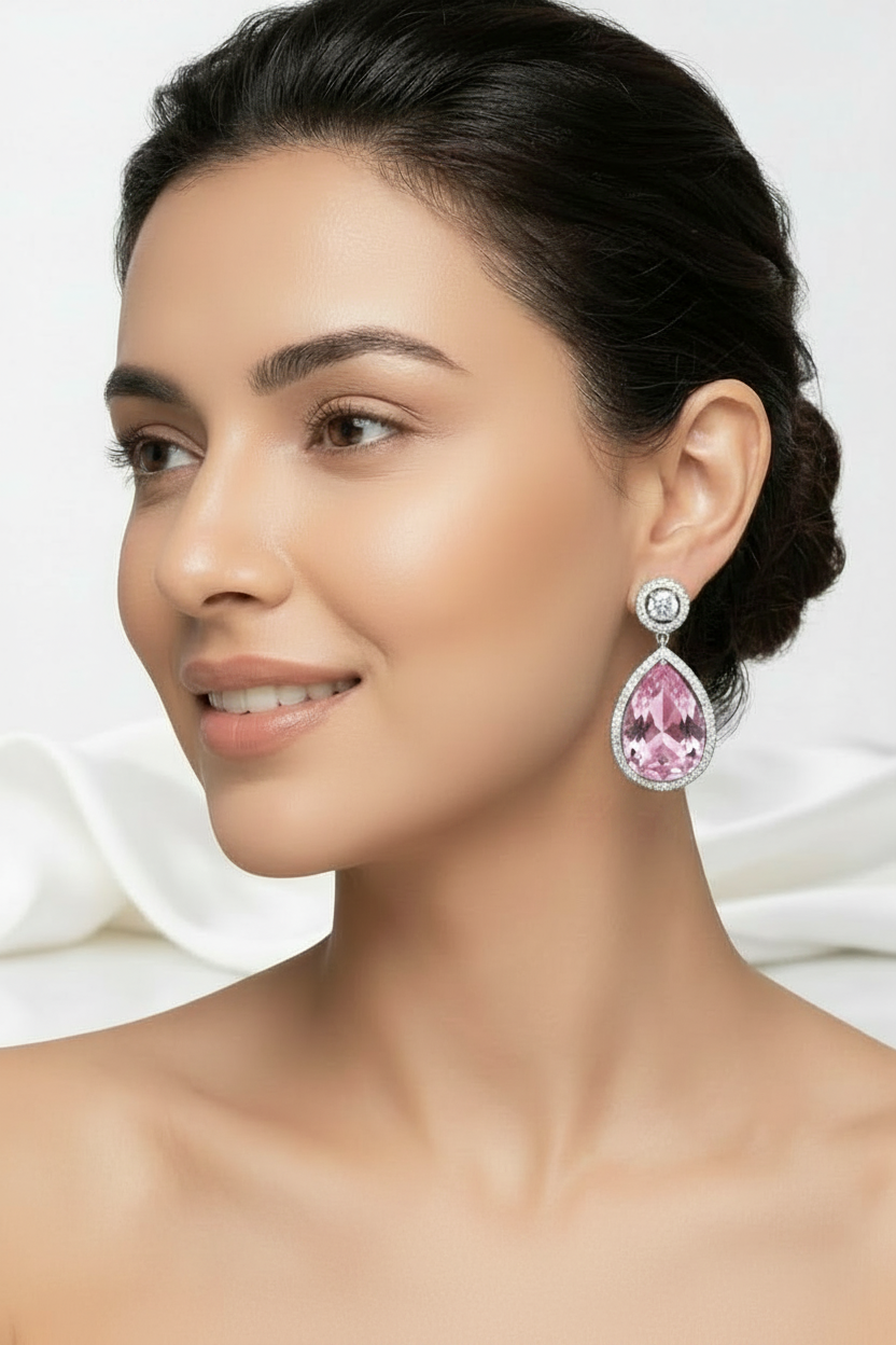 A close-up portrait of a woman wearing a pair of dramatic teardrop dangle earrings with baby pink stones and silver settings, against a soft grey studio background.