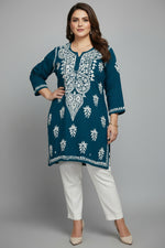 A deep dark teal colour code is #215D6E long-sleeve tunic top with intricate white embroidery, laid flat on a grey herringbone wood floor. The top features a V-neckline with detailed white embroidery, matching embroidered motifs on the front, and border detailing on the cuffs and hem.