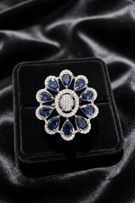 A floral-shaped silver ring with deep blue teardrop gemstones and a central oval cluster of small clear stones, displayed on a black velvet jewelry box over black satin fabric.