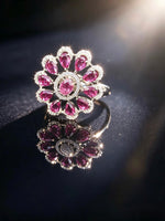 A floral ring featuring pink gemstones and clear accent stones in a silver-toned setting, displayed on a dark reflective surface with a mirrored reflection.