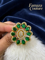 A gold-tone brooch finger ring featuring green teardrop-shaped gemstones and a central cluster of clear stones, held by a hand against a dark blue velvet background with a white faux fur surface. The brand name 'Famaza Couture' is visible in the top right corner.
