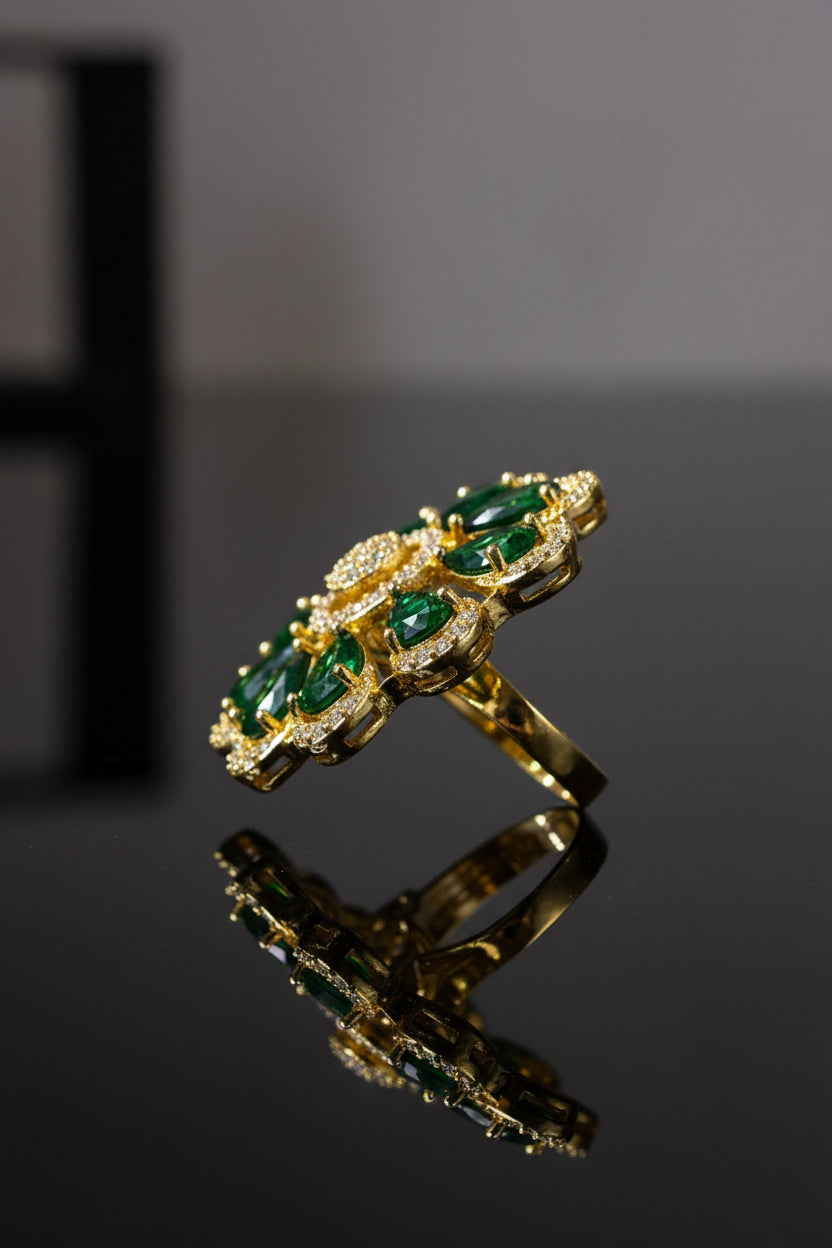A gold-tone finger ring brooch featuring green teardrop-shaped gemstones and a clear crystal centre, displayed on a white faux fur surface.