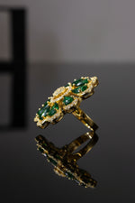 A gold-tone finger ring brooch featuring green teardrop-shaped gemstones and a clear crystal centre, displayed on a white faux fur surface.