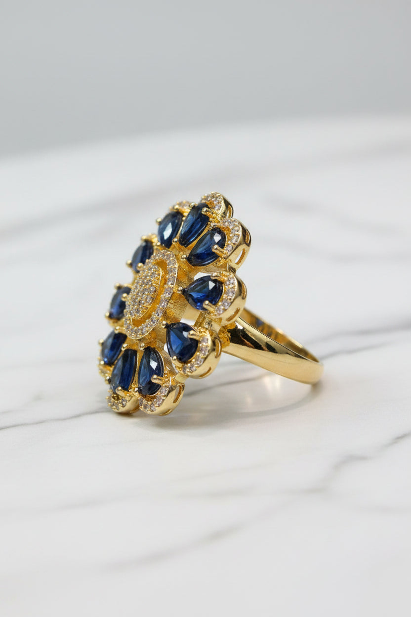 A gold-tone flower-shaped finger ring brooch with deep blue teardrop gemstones and a clear crystal centre, resting on a white textured faux fur surface.