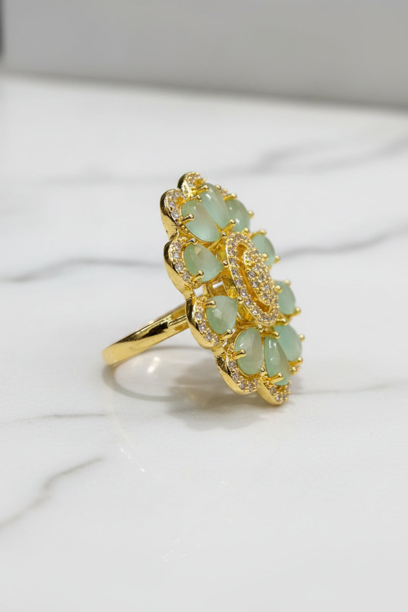 A gold-tone flower-shaped finger ring brooch with pale green teardrop-shaped stones and a textured gold centre, resting on a white faux fur surface.