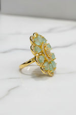 A gold-tone flower-shaped finger ring brooch with pale green teardrop-shaped stones and a textured gold centre, resting on a white faux fur surface.